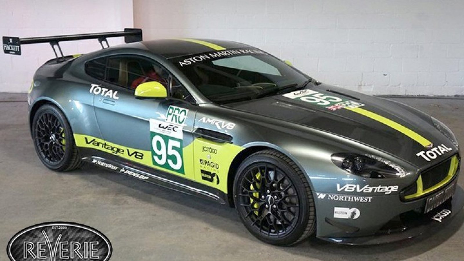 Auto deportivo Aston Martin Vantage en un garaje. El auto es gris con detalles en verde y amarillo, con varios logotipos de patrocinadores. El número "95" está visible en la puerta lateral y en el capó.