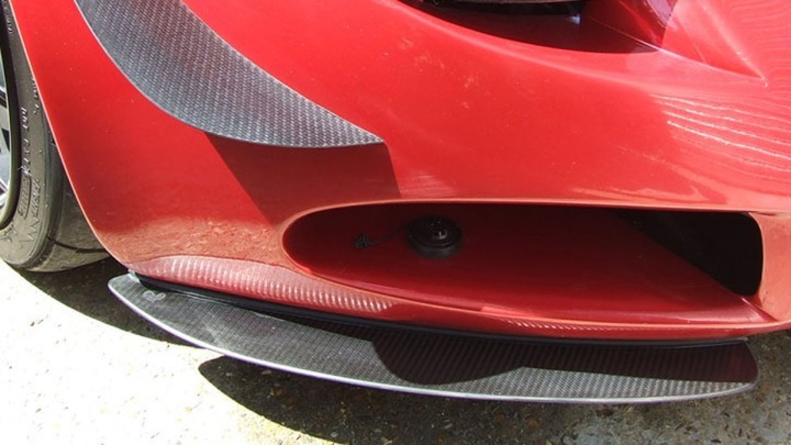 Primer plano de la parte frontal de un automóvil rojo deportivo. Se observa el detalle de la carrocería en fibra de carbono alrededor de la defensa y una entrada de aire en el parachoques inferior.